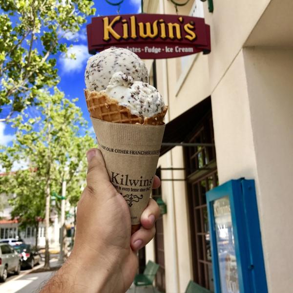 A hand holds a double-scoop waffle cone of cookies-and-cream ice cream in front of a Kilwins storefront on a sunny day.