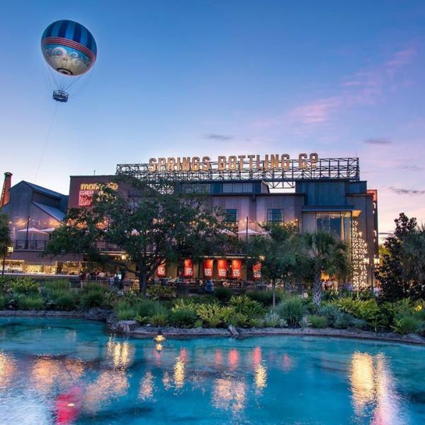 Evening view of Disney Springs with the iconic Aerophile balloon floating above the Springs Bottling Co. building, surrounded by palm trees and water reflecting the sunset sky.