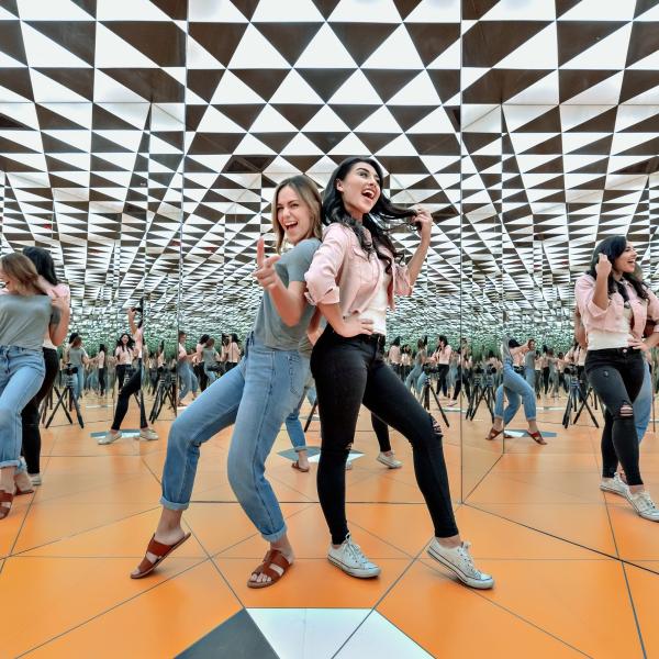 Two women pose playfully inside the Museum of Illusions, surrounded by mirrored walls and a geometric black-and-white ceiling that creates endless reflections.