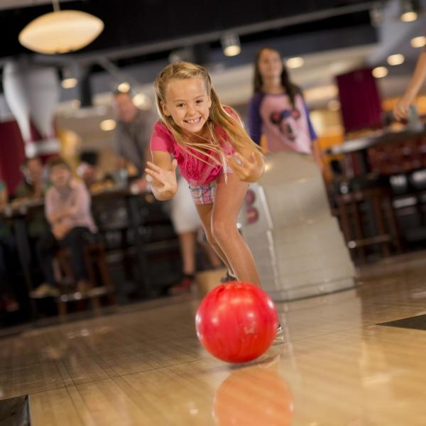 Smiling young girl rolls a red bowling ball down the lane while friends and family cheer her on in the background.