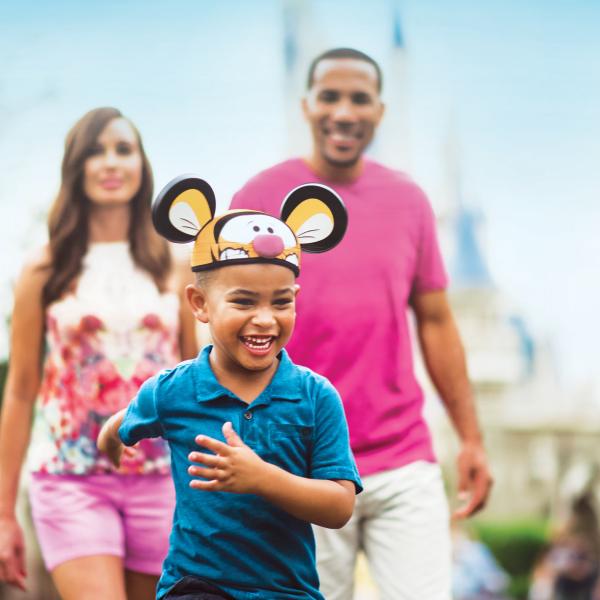 Smiling young boy wearing themed mouse ears runs ahead of his parents at a theme park in Kissimmee, Florida.