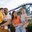 A family of four smiles and poses together in front of a looping roller coaster at a theme park, with parents holding their two young children.